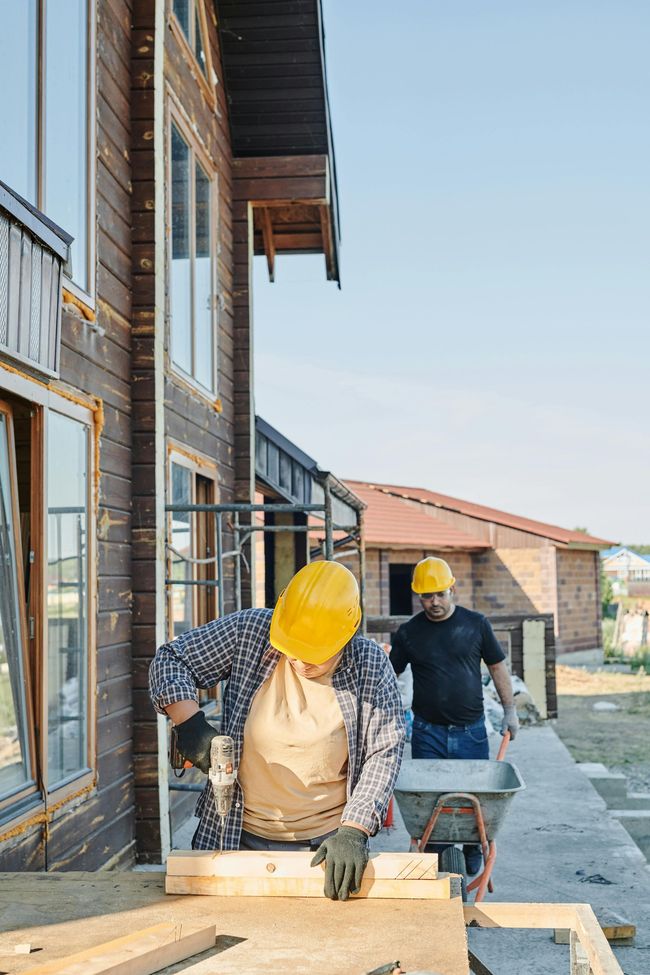 Two construction workers in hard hats work on the exterior of a brown wooden house.
