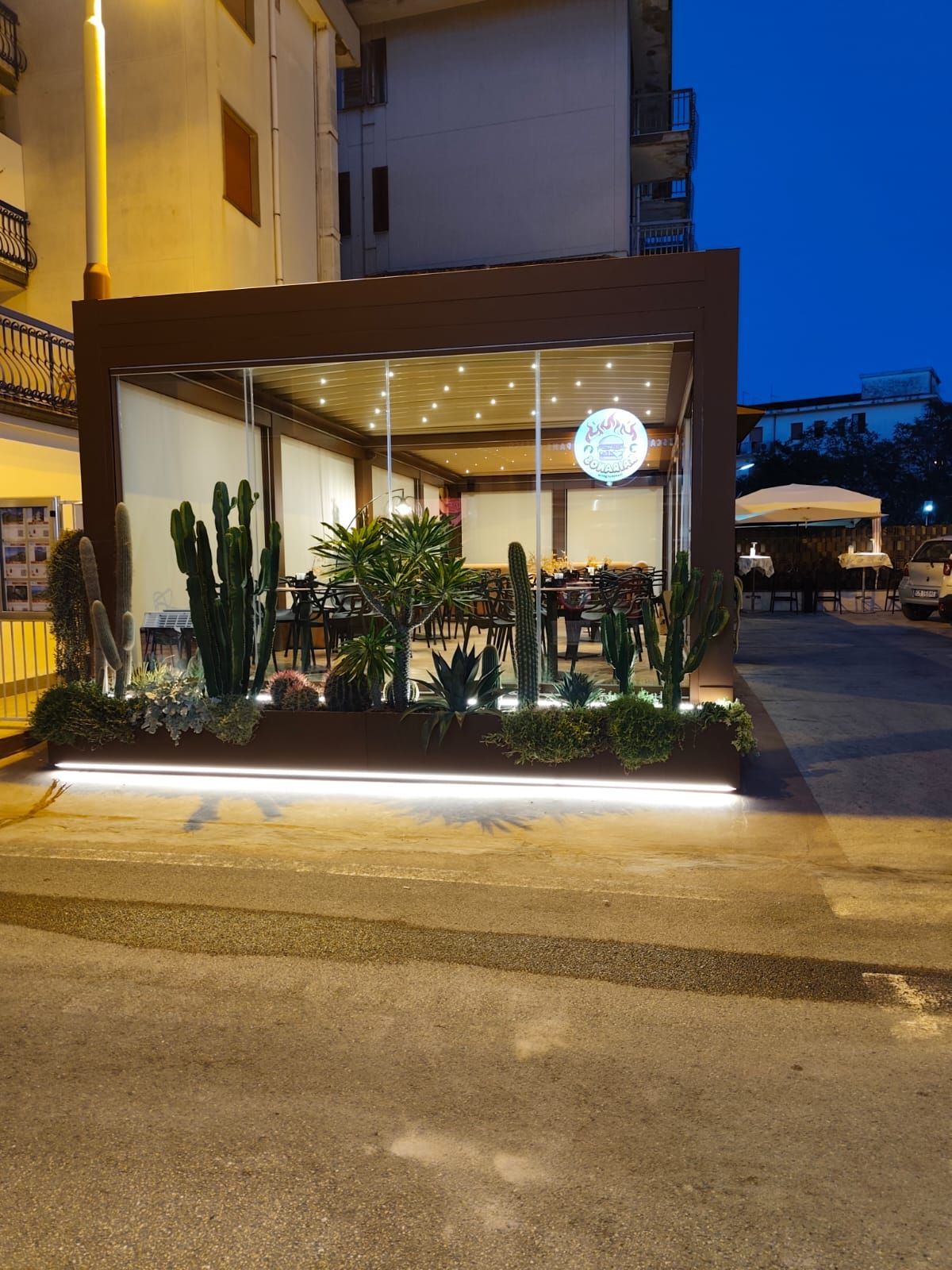 Ristorante con pareti in vetro, cactus e perimetro illuminato. Ambiente notturno con tavoli visibili all'interno.