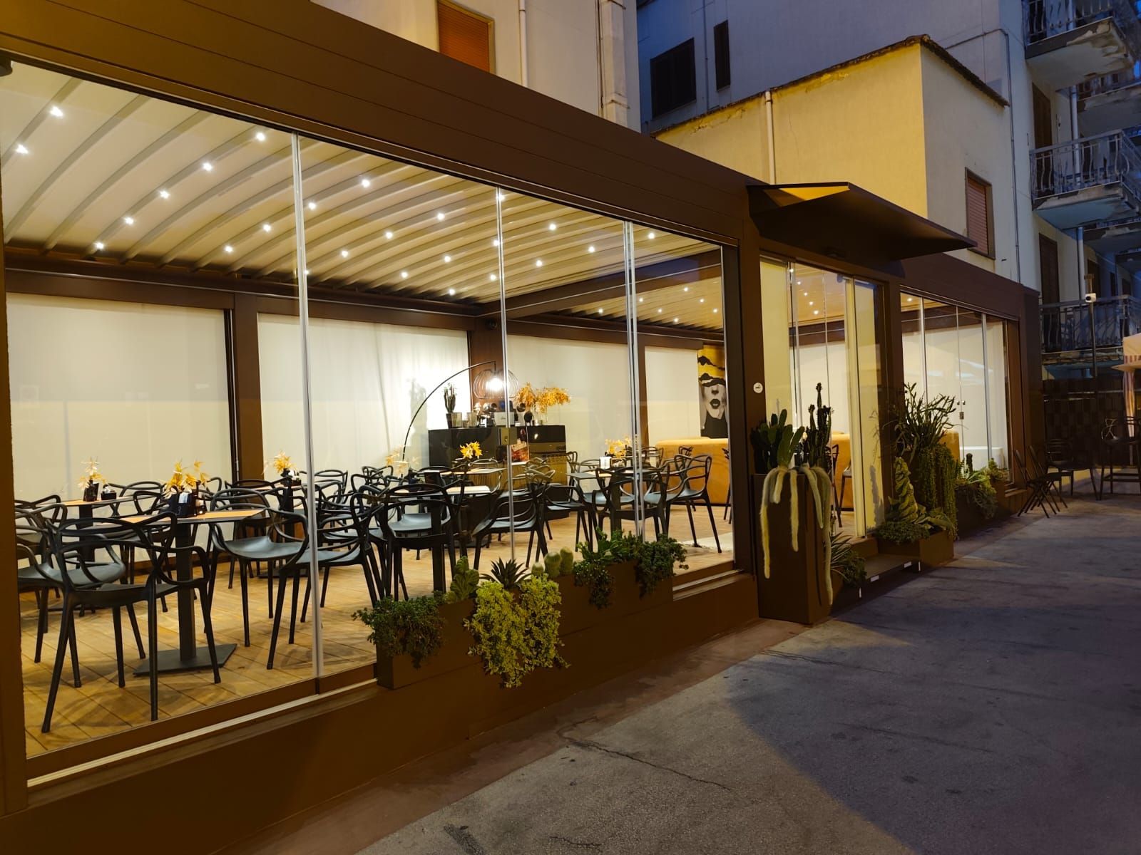 Patio del ristorante con tavoli, sedie e piante dietro pareti di vetro sotto una tenda da sole marrone.