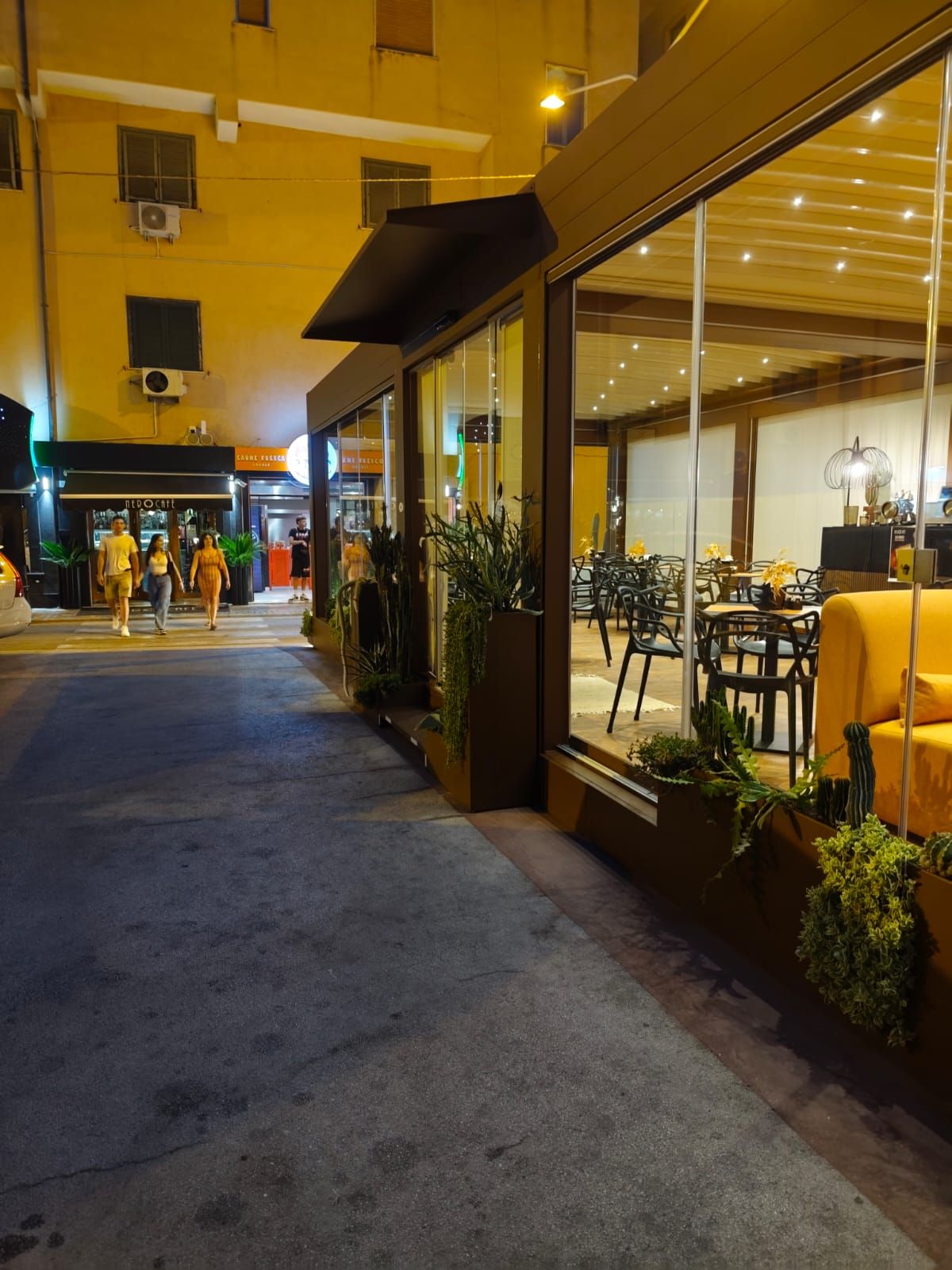 Esterno del ristorante di notte. Pareti in vetro, edificio giallo, gente che cammina per strada.