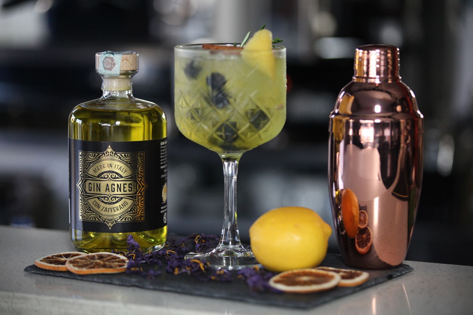 Cocktail con bottiglia, shaker, limone e fette di arancia essiccate su un bancone.