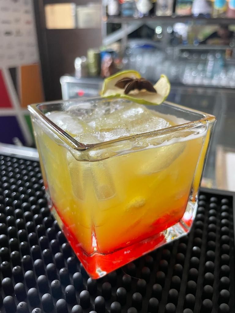 Cocktail in un bicchiere quadrato, con strati di rosso e arancione, guarnito con ghiaccio e frutta verde su un bancone.