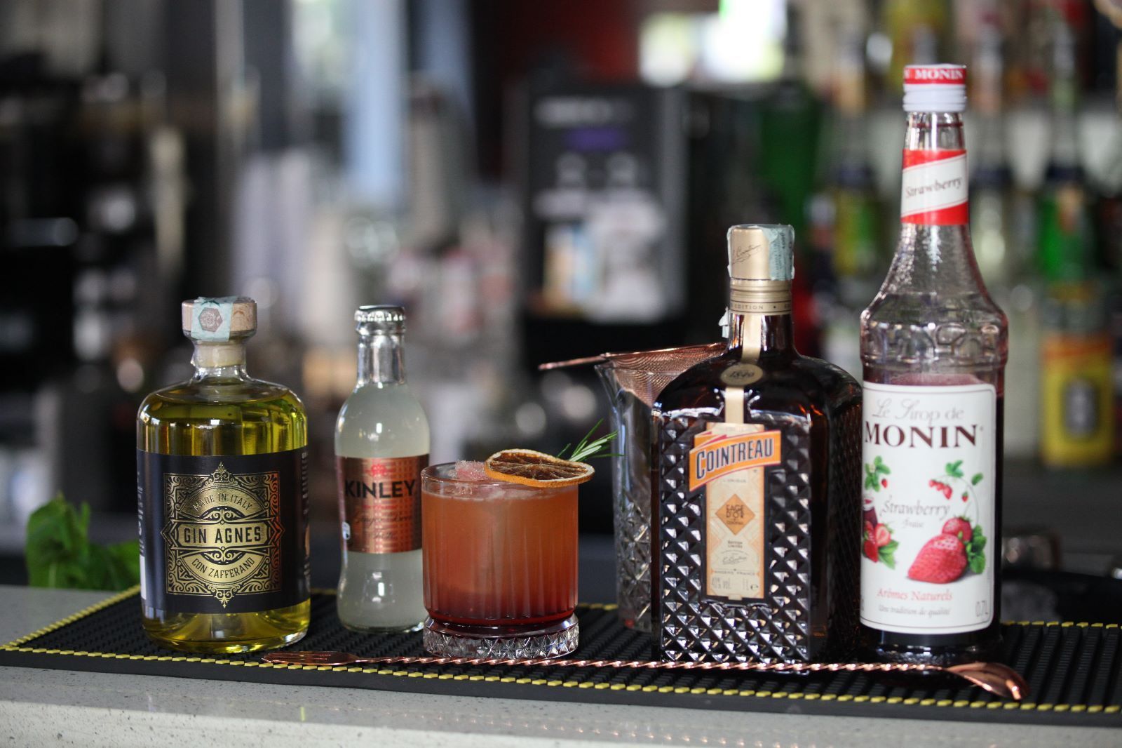 Cocktail servito al bancone con bottiglie di liquore e sciroppi. Il drink è guarnito con scorza d'arancia.