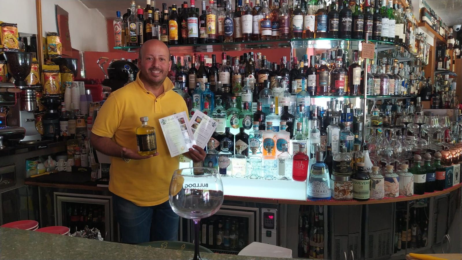 Un uomo con una maglietta gialla tiene in mano dei drink, in piedi al bar, circondato da bottiglie di alcolici.