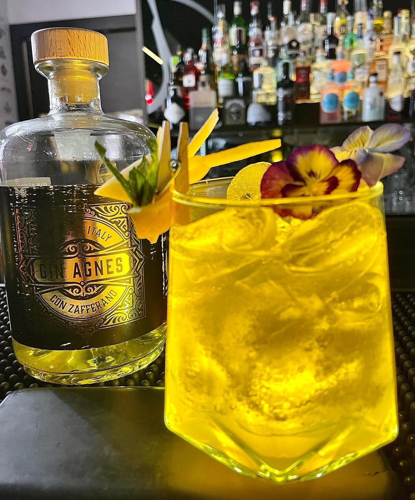 Cocktail giallo in un bicchiere di cristallo, guarnito con fiori e agrumi, accanto a una bottiglia di liquore.