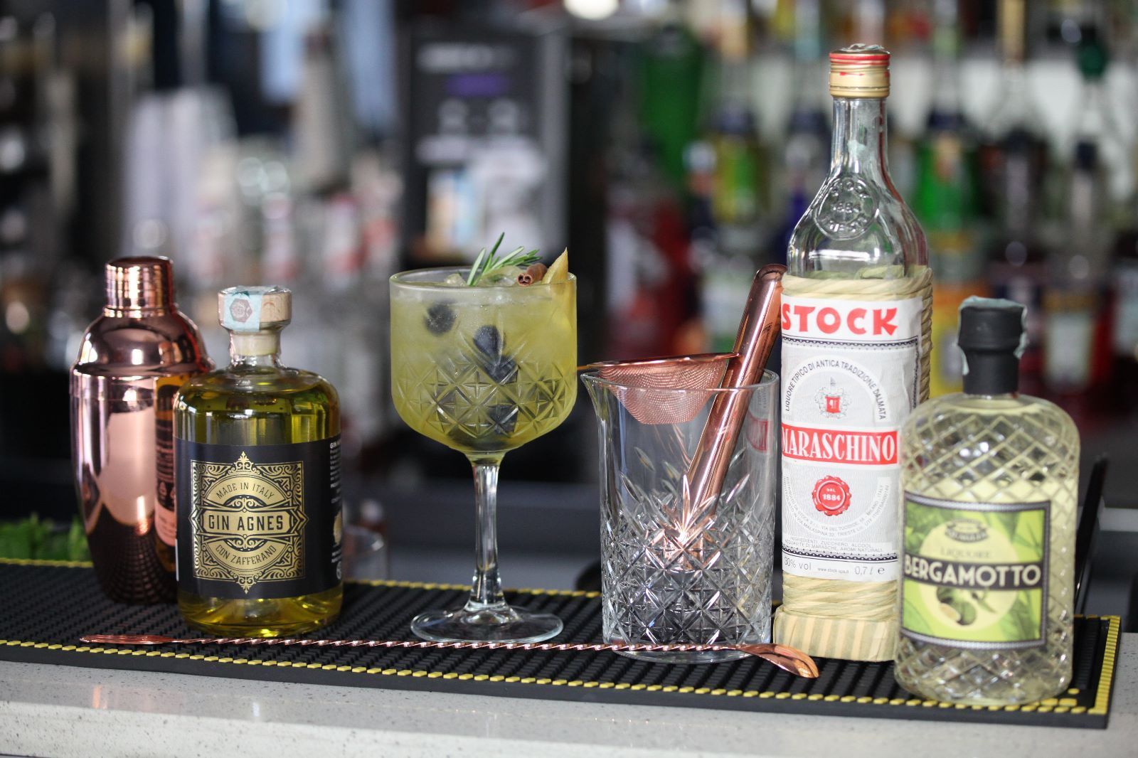 Allestimento per cocktail con bottiglie di liquore, uno shaker, un drink miscelato, un mixing glass e un cucchiaio per mescolare su un bancone.