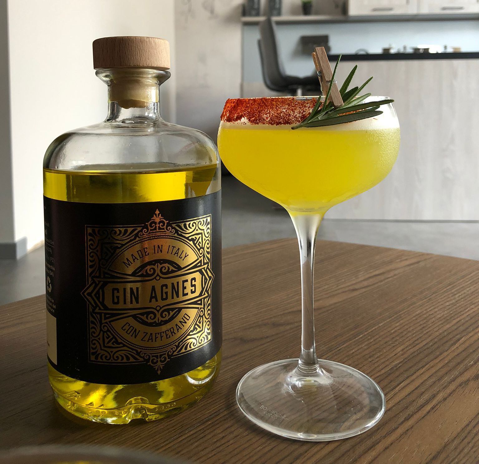 Bottiglia di Gin Agnes accanto a un cocktail giallo con guarnizione di rosmarino e bordo di zucchero rosso su una superficie di legno.
