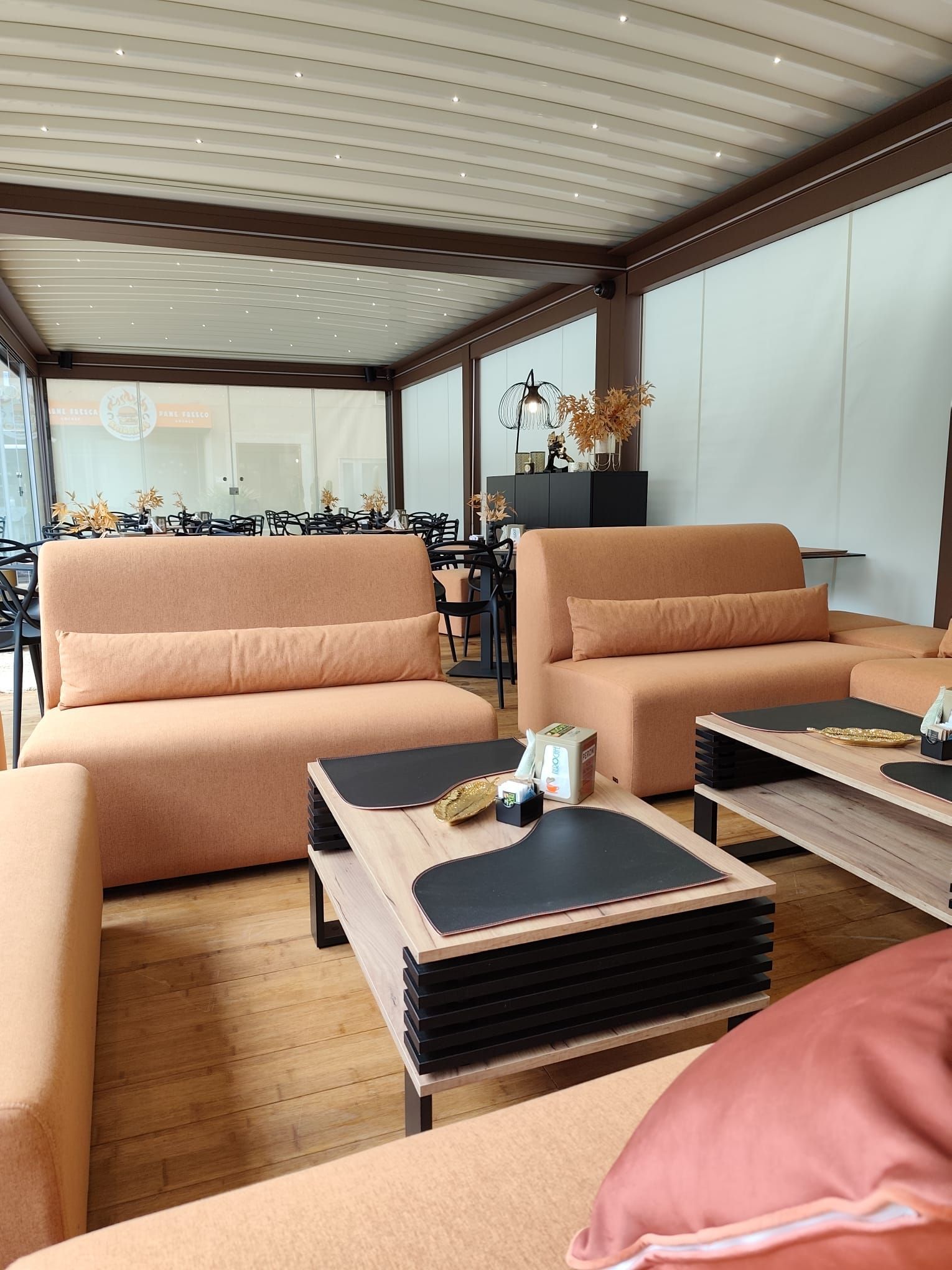 Area salotto all'aperto con divani arancioni, tavoli neri e tetto retrattile.