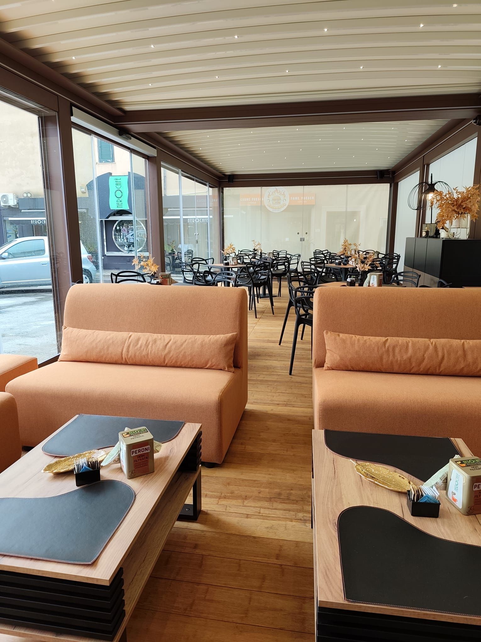 Area esterna del ristorante con divani arancioni, tavoli e sedie nere. Pavimento e soffitto in legno.