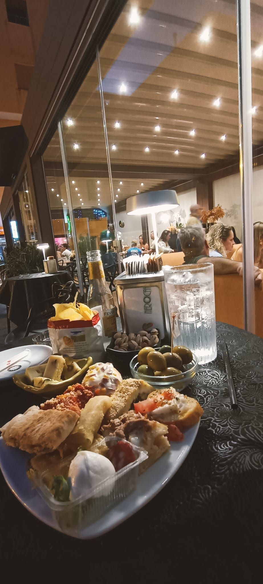 Un tavolo con antipasti, bevande e l'interno sfocato del ristorante visibile attraverso una parete di vetro.