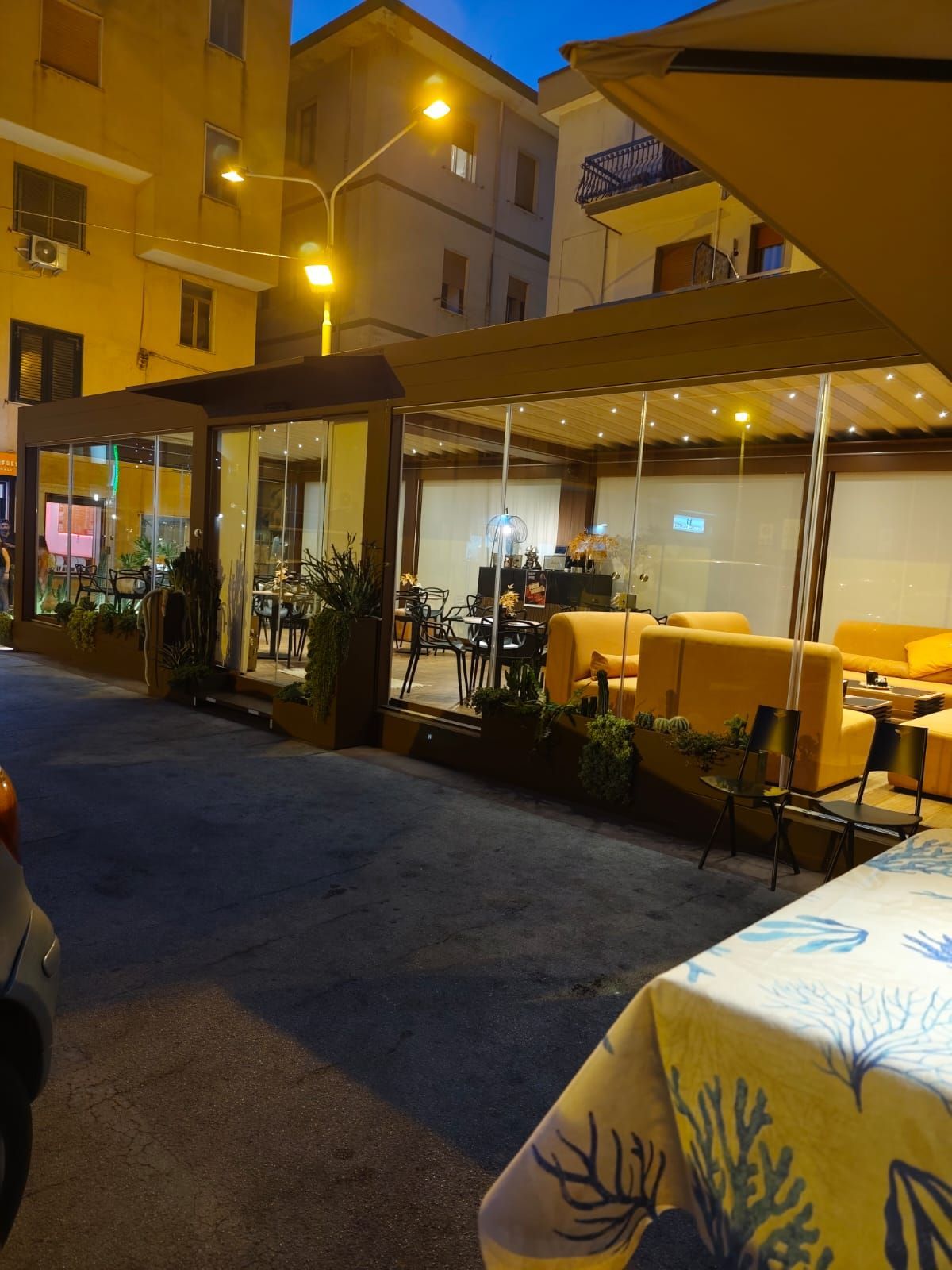 Ristorante all'aperto di notte con pareti in vetro, divani gialli, tavoli, lampioni ed edifici sullo sfondo.