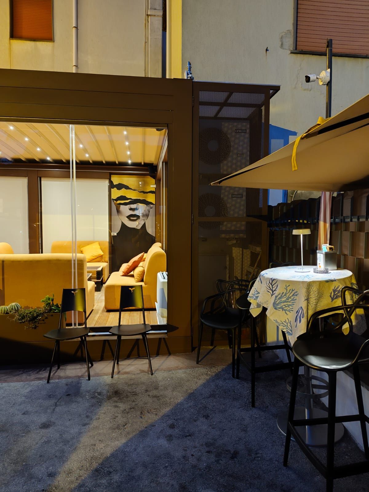 Posti a sedere all'aperto del bar: divano giallo dietro vetro, tavoli, sedie nere, opere d'arte, ombrellone da giardino, di notte.