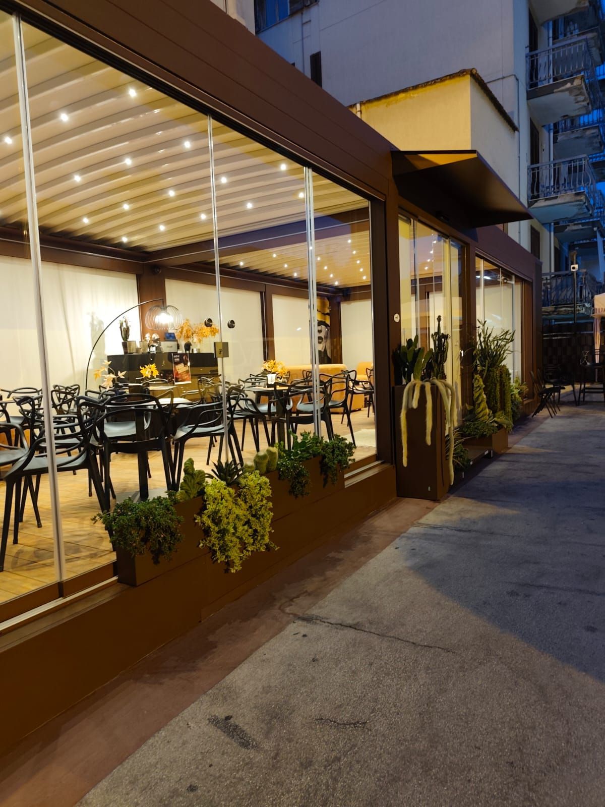 Ristorante all'aperto con pareti in vetro, tavoli e sedie. Piante in fioriere costeggiano l'esterno.