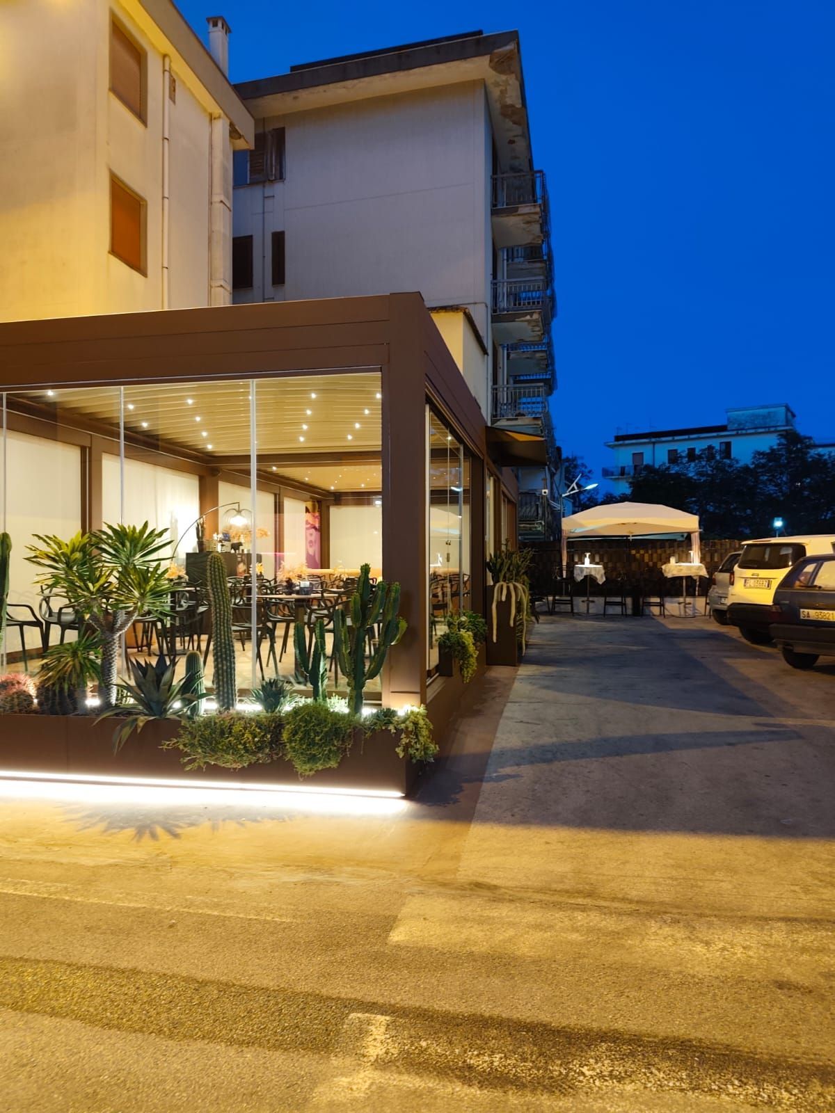 Ristorante all'aperto con pareti in vetro, tenda da sole marrone e piante al tramonto. Sono visibili auto e un edificio.