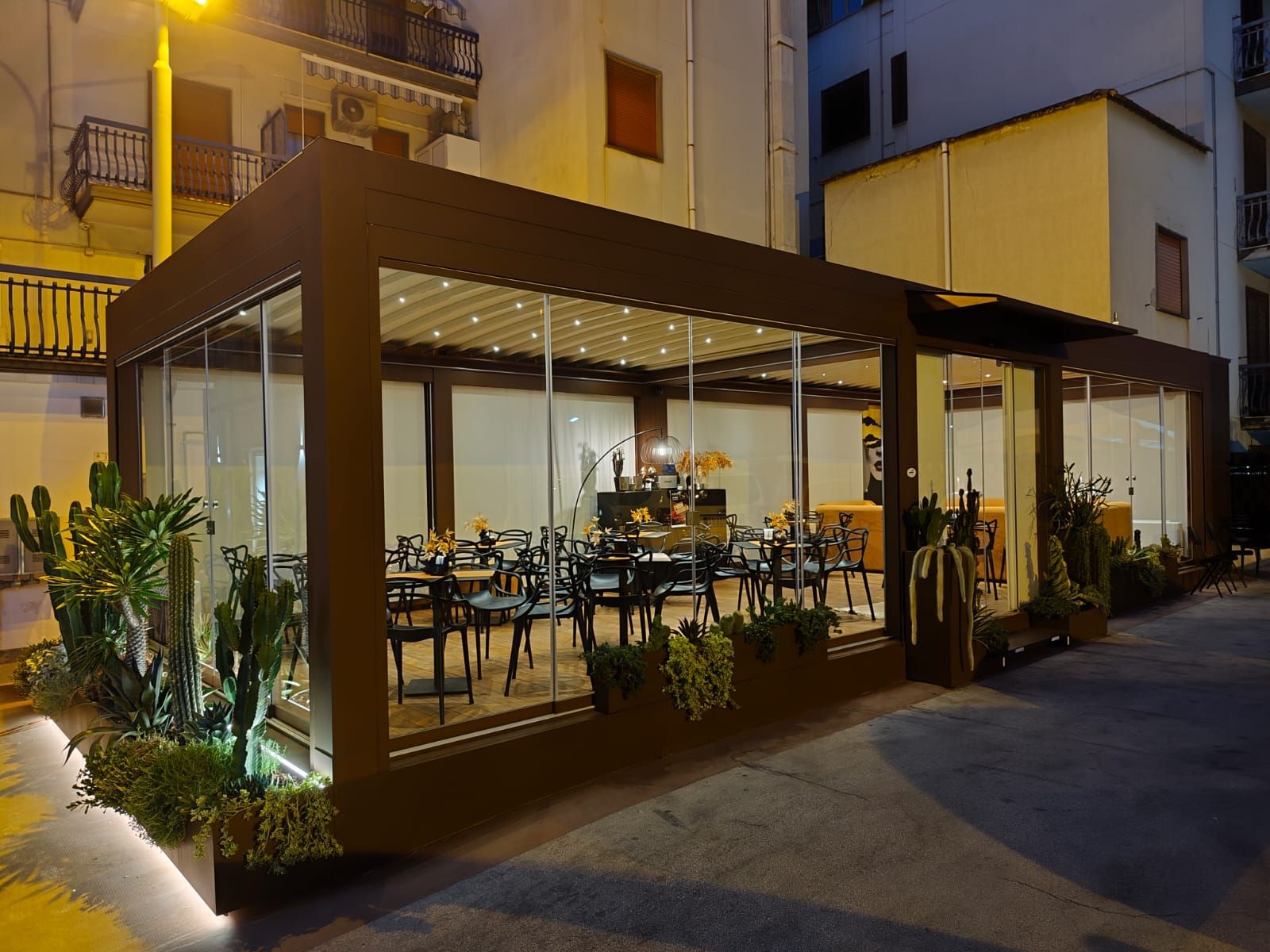 Patio del ristorante con pareti in vetro, struttura marrone, tavoli, sedie e piante; illuminato di notte.