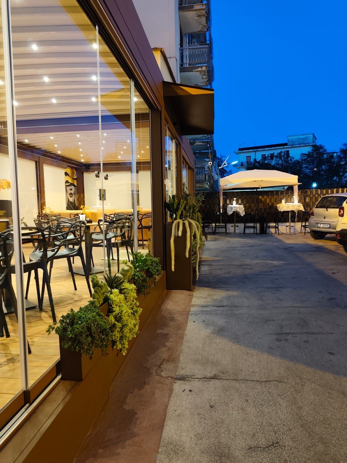 Vista esterna di un ristorante con vetrate, tavoli, sedie e piante. Scenario serale con cielo azzurro.