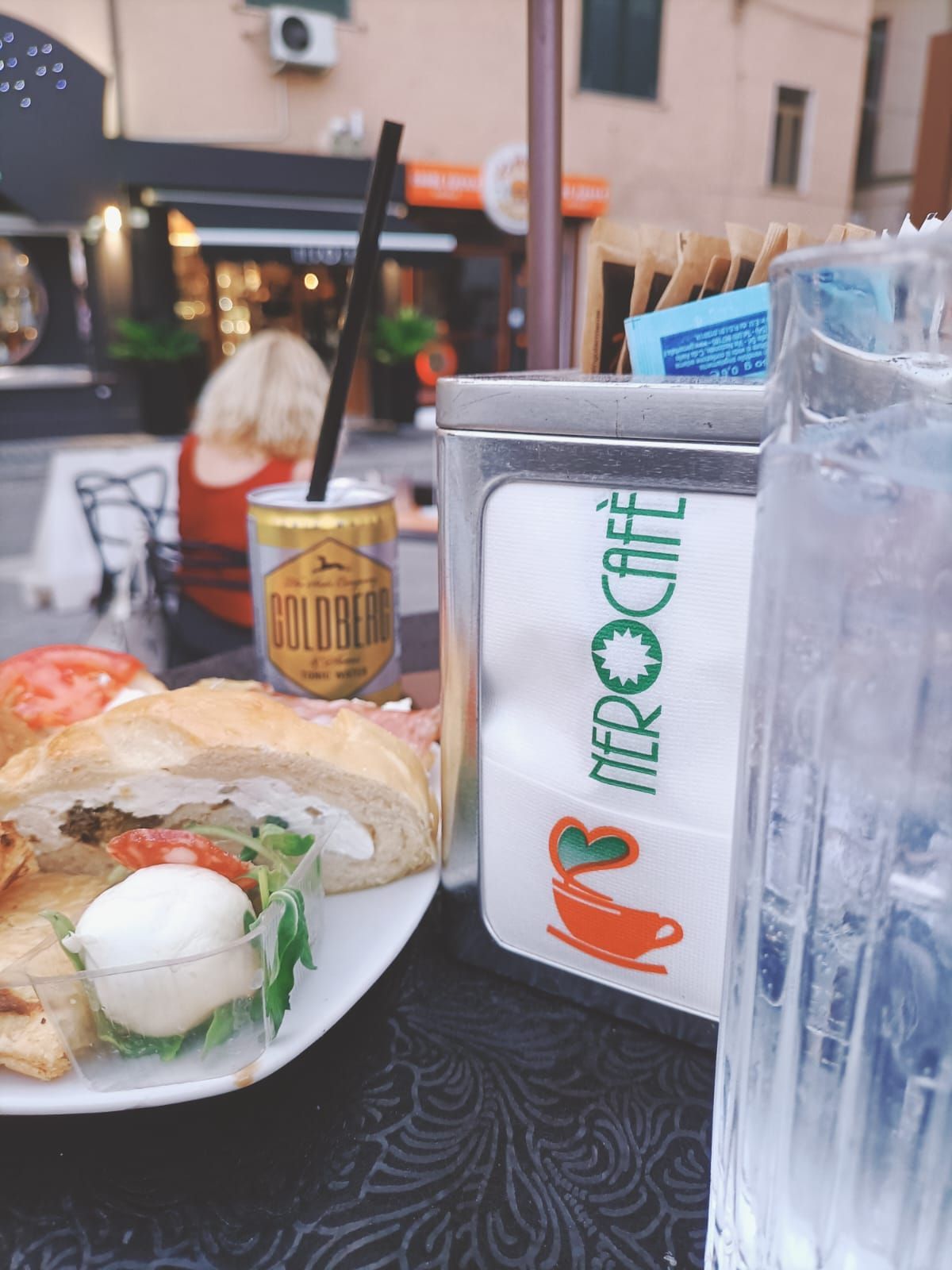 Tavolo con cibo e bevande in un bar all'aperto. Portatovaglioli, persona sullo sfondo.