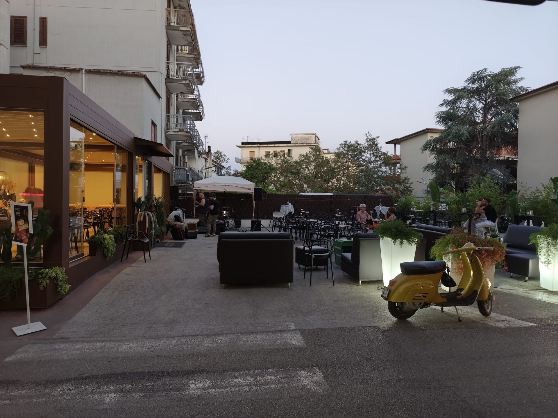 Scena di un bar all'aperto con scooter giallo, tavoli e clienti. Edifici sullo sfondo.