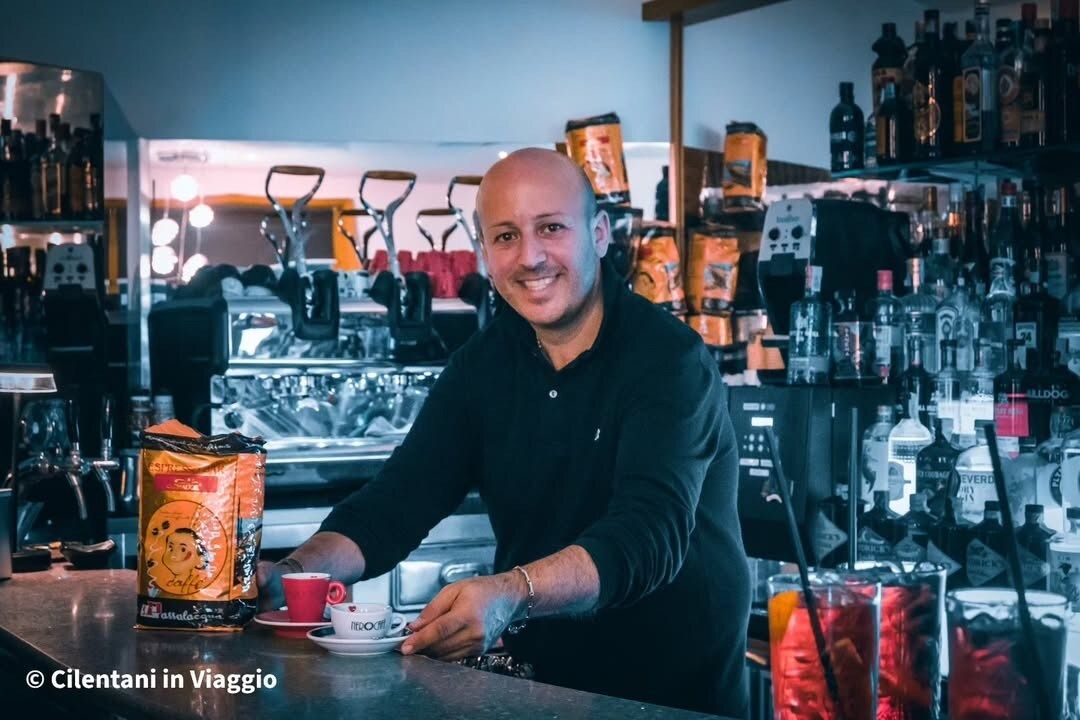 Uomo sorridente dietro un bancone, con in mano una tazza di caffè. Sullo sfondo, bottiglie e macchina per l'espresso.