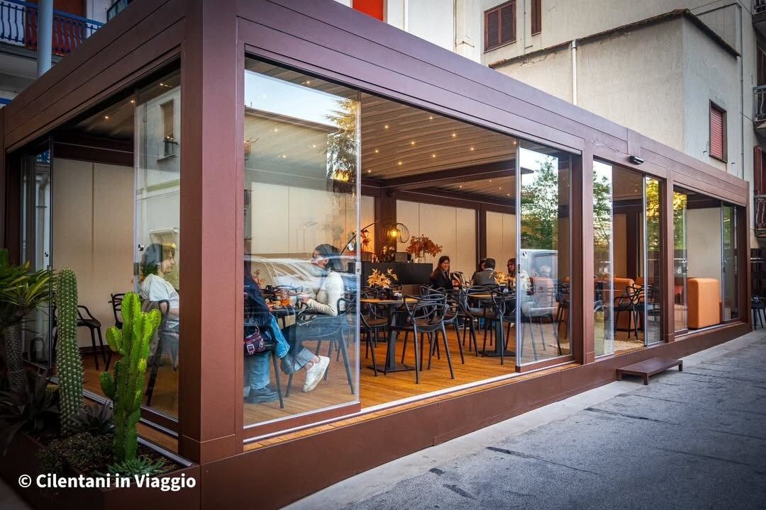 Bar con struttura marrone, ampie finestre, tavoli e persone che cenano all'interno. All'esterno si vedono piante verdi.