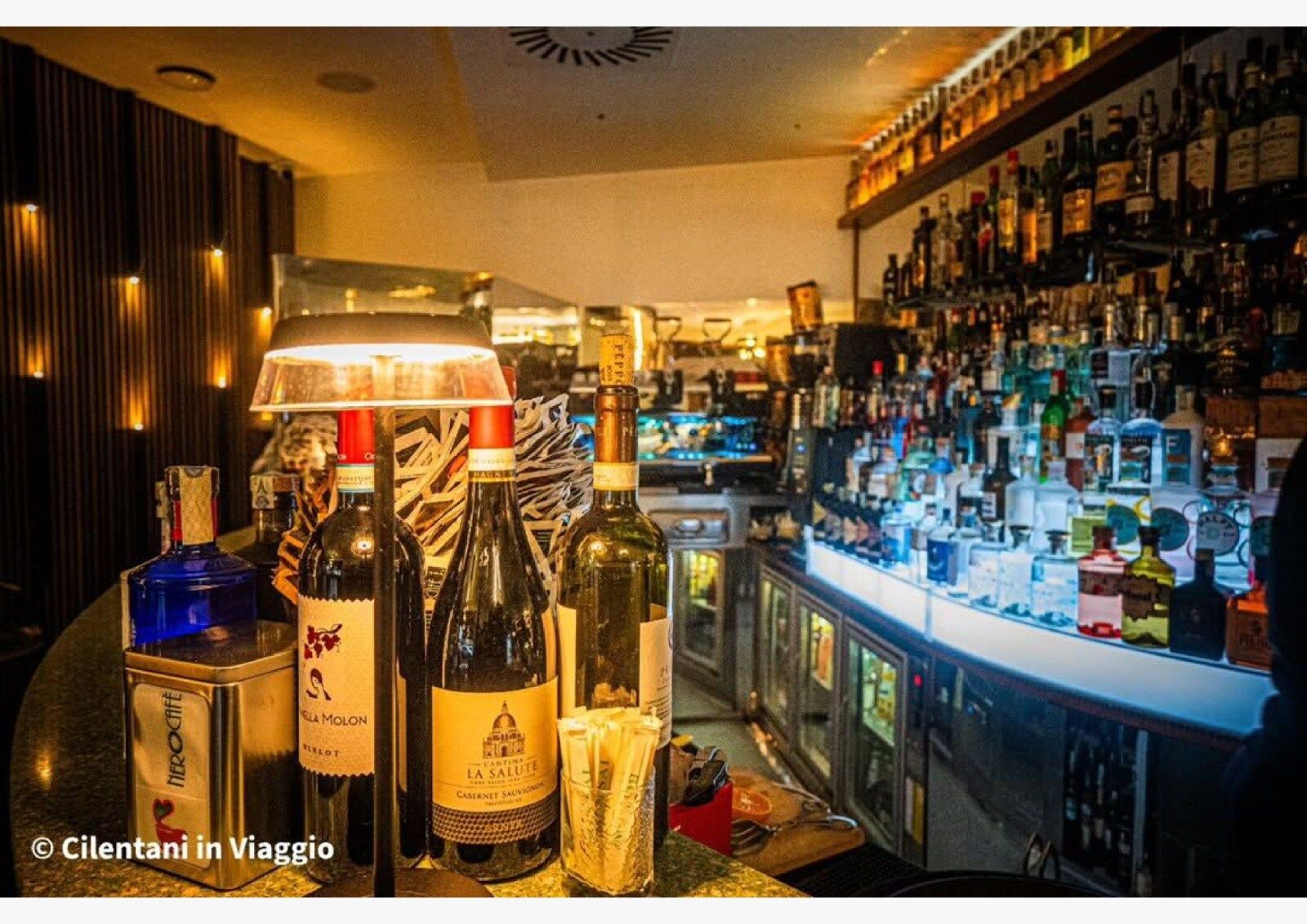 Bar interno con bottiglie di liquore sugli scaffali, bottiglie sul bancone e una calda luce.