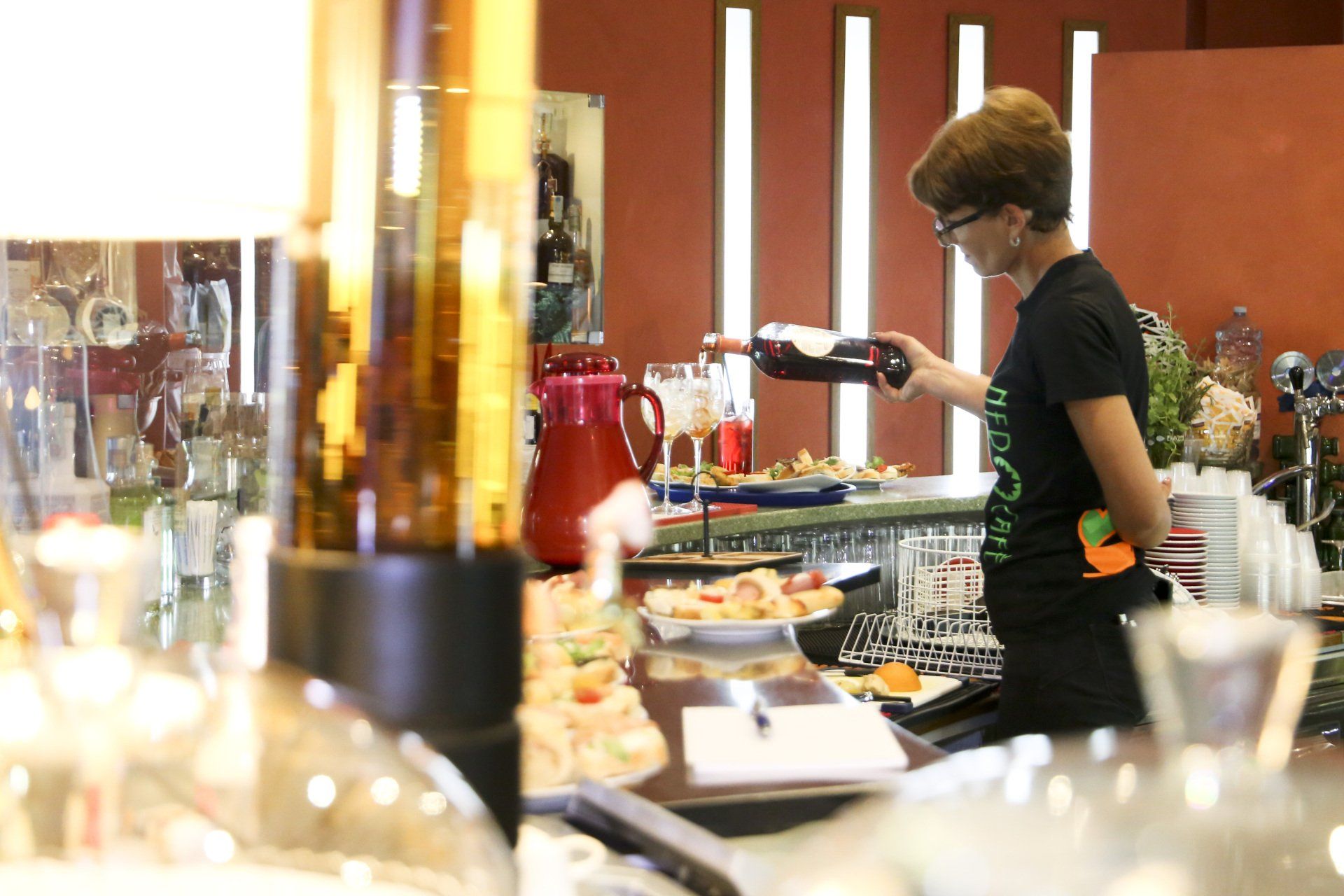 Donna che versa vino dietro un bancone, dove si vedono cibo e bicchieri.