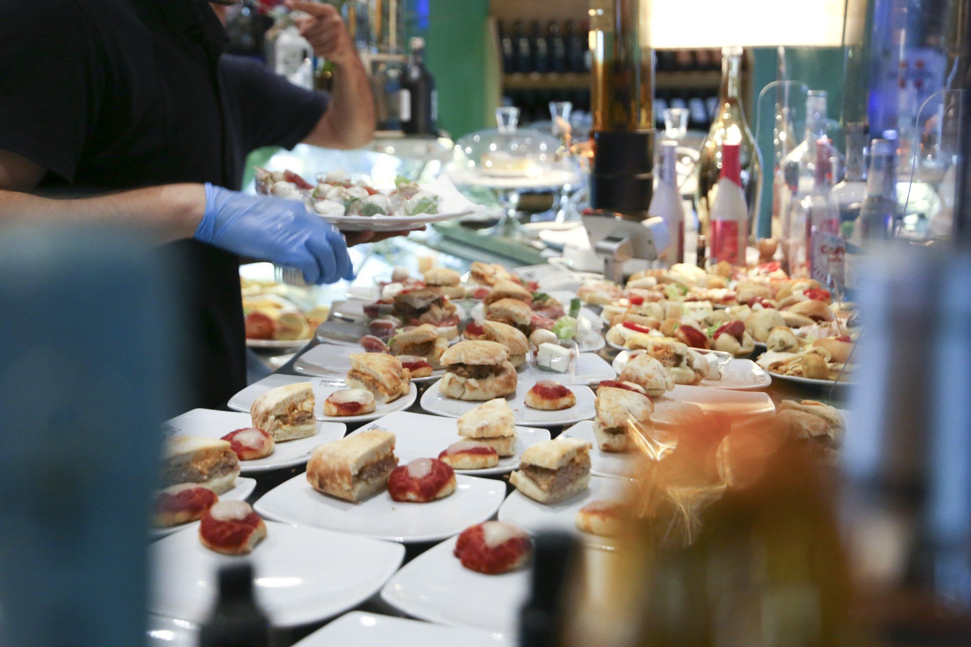 Un buffet con piccoli piatti di antipasti, una persona che indossa guanti blu serve il cibo.
