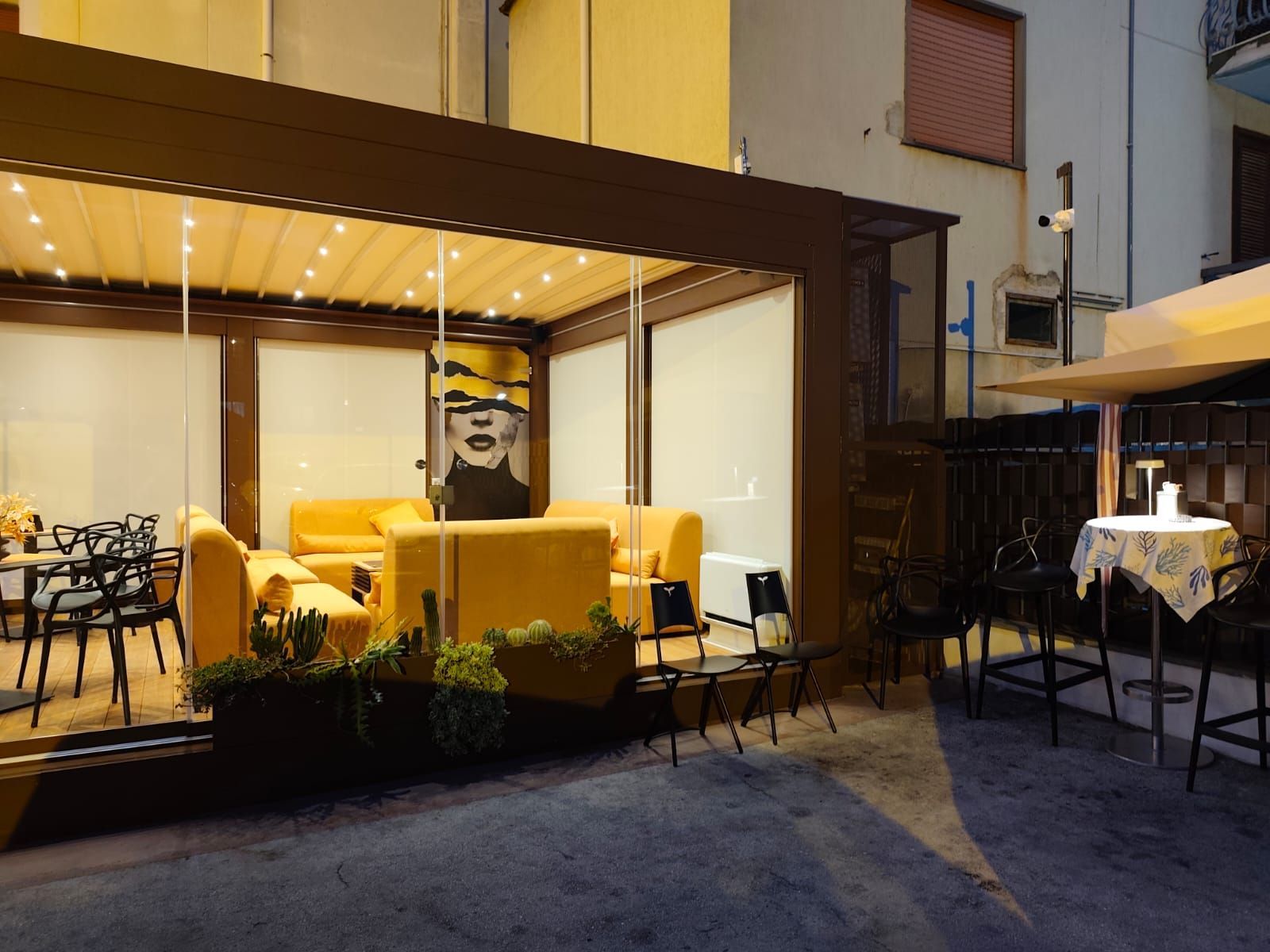 Bar all'aperto con posti a sedere gialli, pareti in vetro e una tenda da sole.
