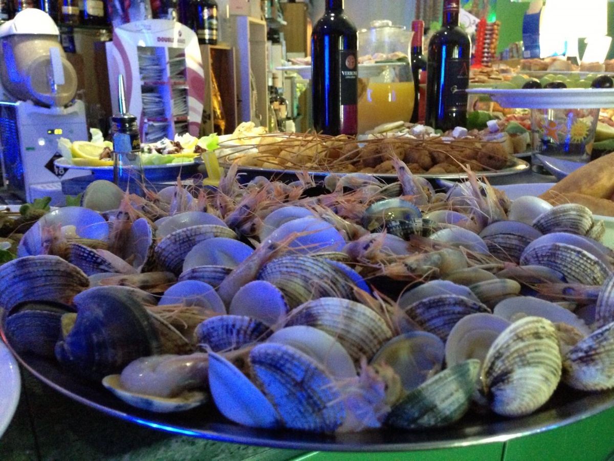 Buffet di pesce: vongole e gamberi su un vassoio, con bottiglie di vino e altri prodotti alimentari sullo sfondo.