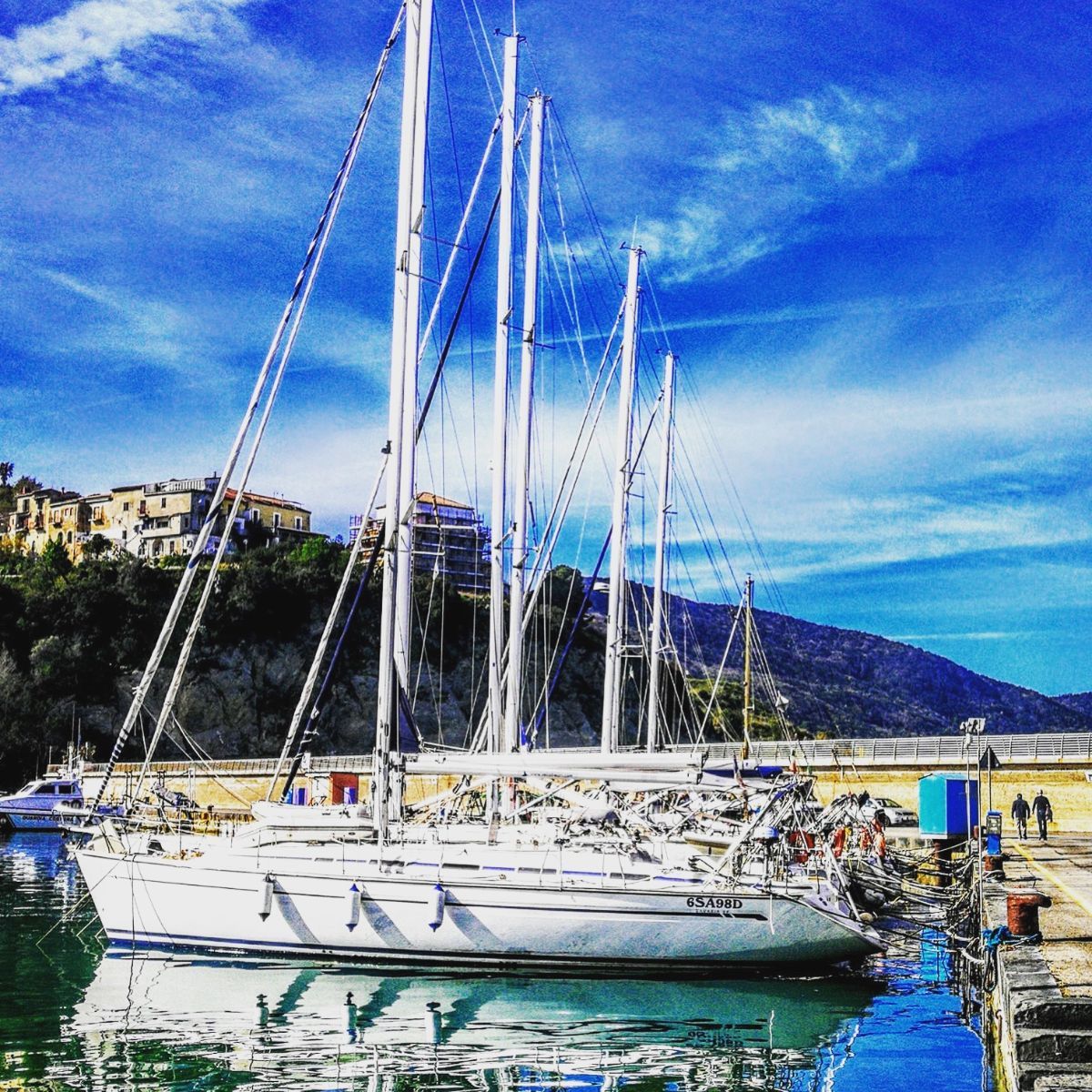 Barche a vela ormeggiate nel porto, alberi contro il cielo azzurro, edifici su una scogliera sullo sfondo.