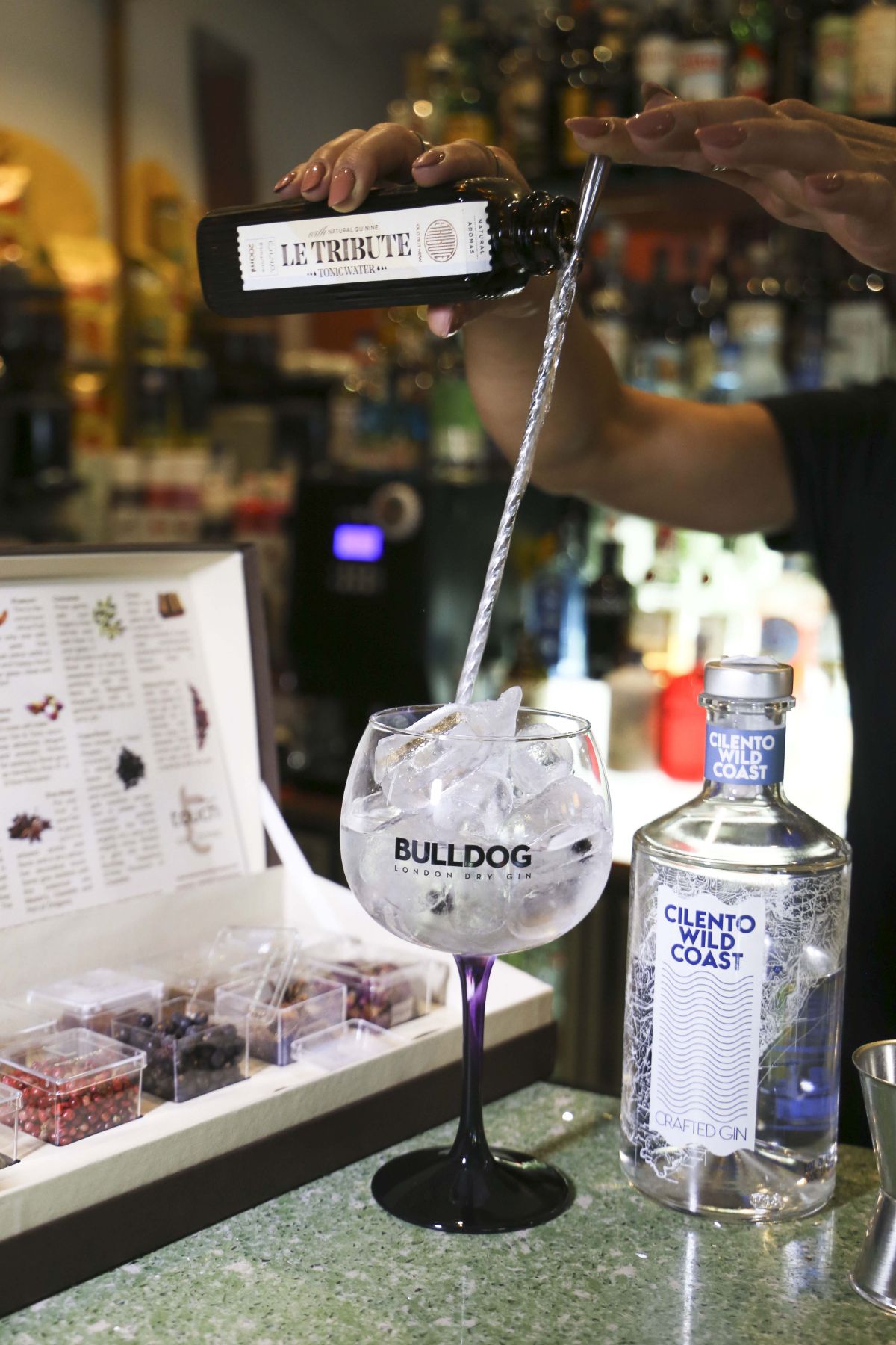 Il barista versa il gin da una bottiglia in un bicchiere pieno di ghiaccio; sono visibili un'altra bottiglia di gin e una scatola di guarnizioni.