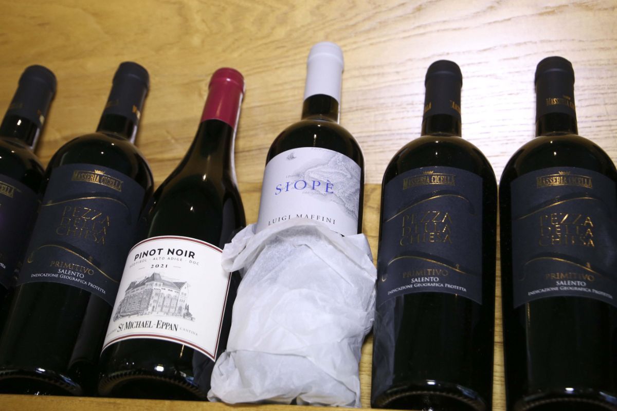 Bottiglie di vino su uno scaffale di legno, alcune con etichette e una avvolta in carta bianca.