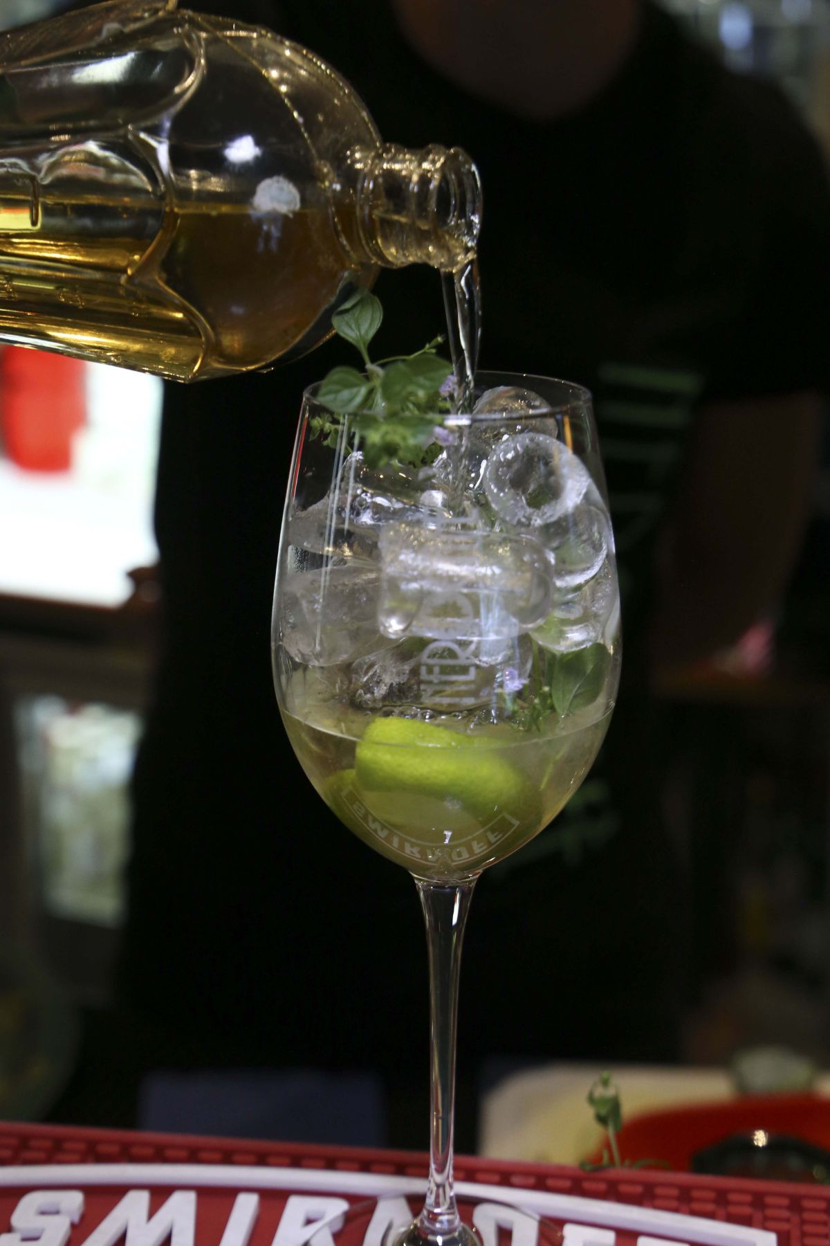 Un barista versa un liquido dorato da una bottiglia in un bicchiere alto con ghiaccio, lime e menta.