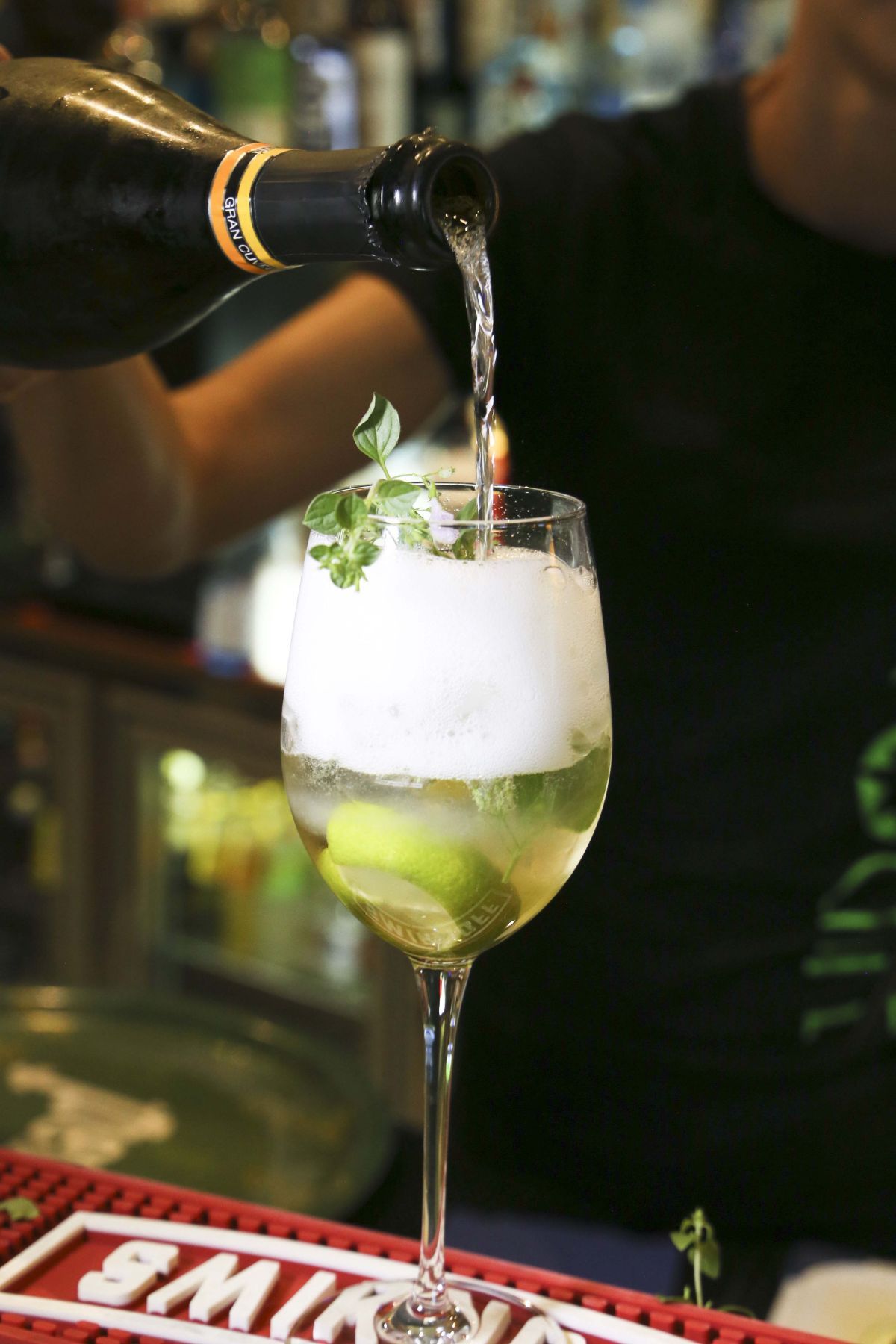 Barista che versa vino spumante in un bicchiere alto con lime, erbe aromatiche e schiuma.