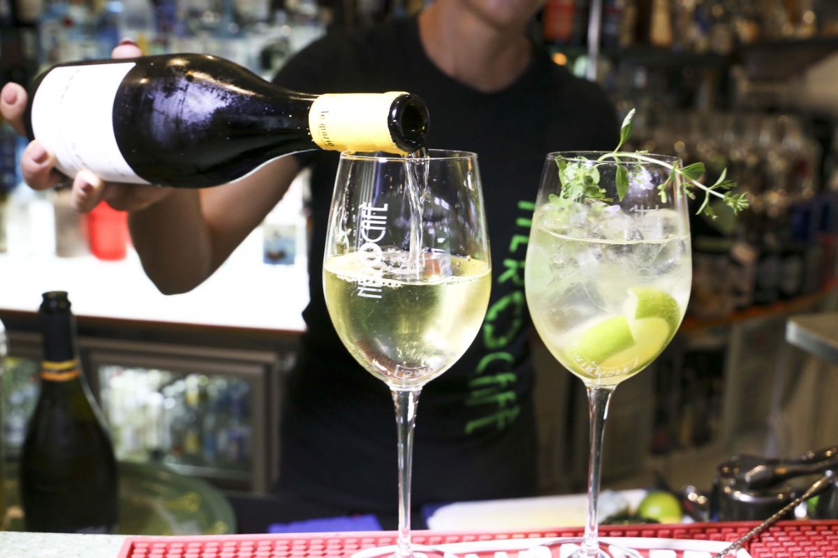 Barista che versa vino bianco in un bicchiere, accanto a un altro bicchiere da cocktail, ambiente da bar.