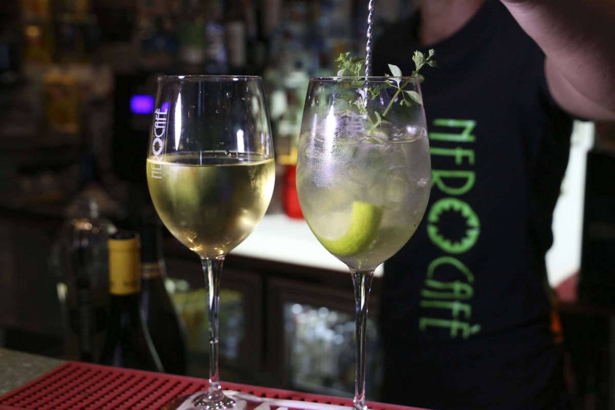 Due bicchieri da vino, uno pieno di vino bianco, l'altro con un cocktail guarnito con lime ed erbe aromatiche; un barista mescola.