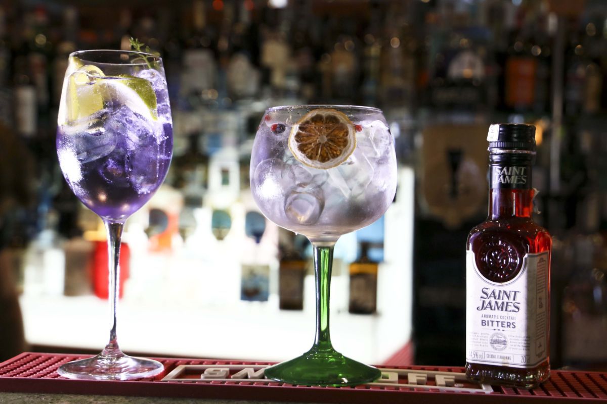Due cocktail a base di gin e una bottiglia di bitter Saint Jame's su un bancone del bar. I cocktail sono viola e trasparenti, con ghiaccio e guarnizioni.