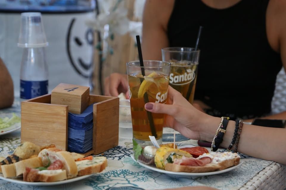 Persona che tiene in mano un bicchiere di tè freddo al tavolo con degli snack e un'altra persona sullo sfondo.