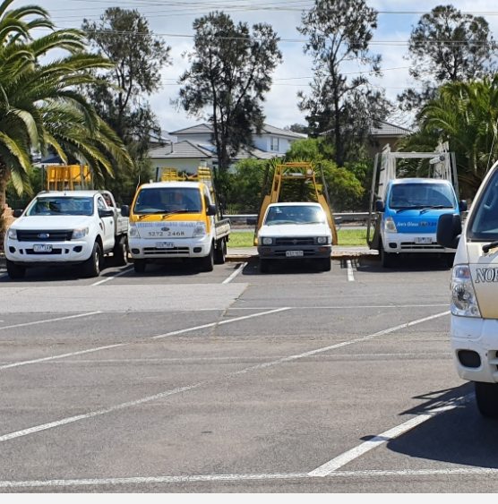 A parking lot with several utility trucks and cars parked in it, with trees and buildings in the background.