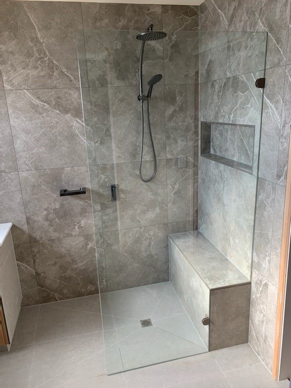 Shower with glass enclosure, gray tiled walls and floor, built-in bench and shower head, stainless steel fixtures.