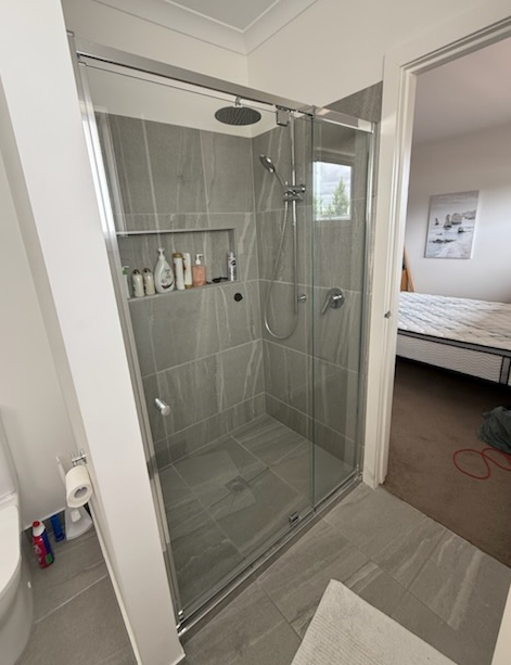 Bathroom with gray tiled shower, glass door, and built-in shelf with toiletries, open doorway leads to a bedroom.