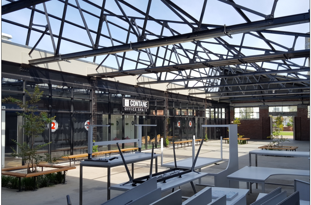 Open-air retail space with black metal framework, several overturned tables and benches, and storefronts.