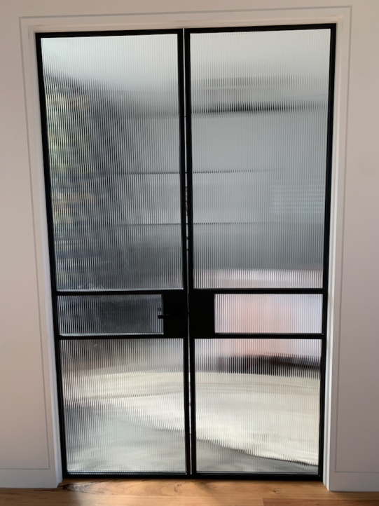 Black-framed glass doors with vertical textured glass panels. The setting is an interior with white walls and a wooden floor.