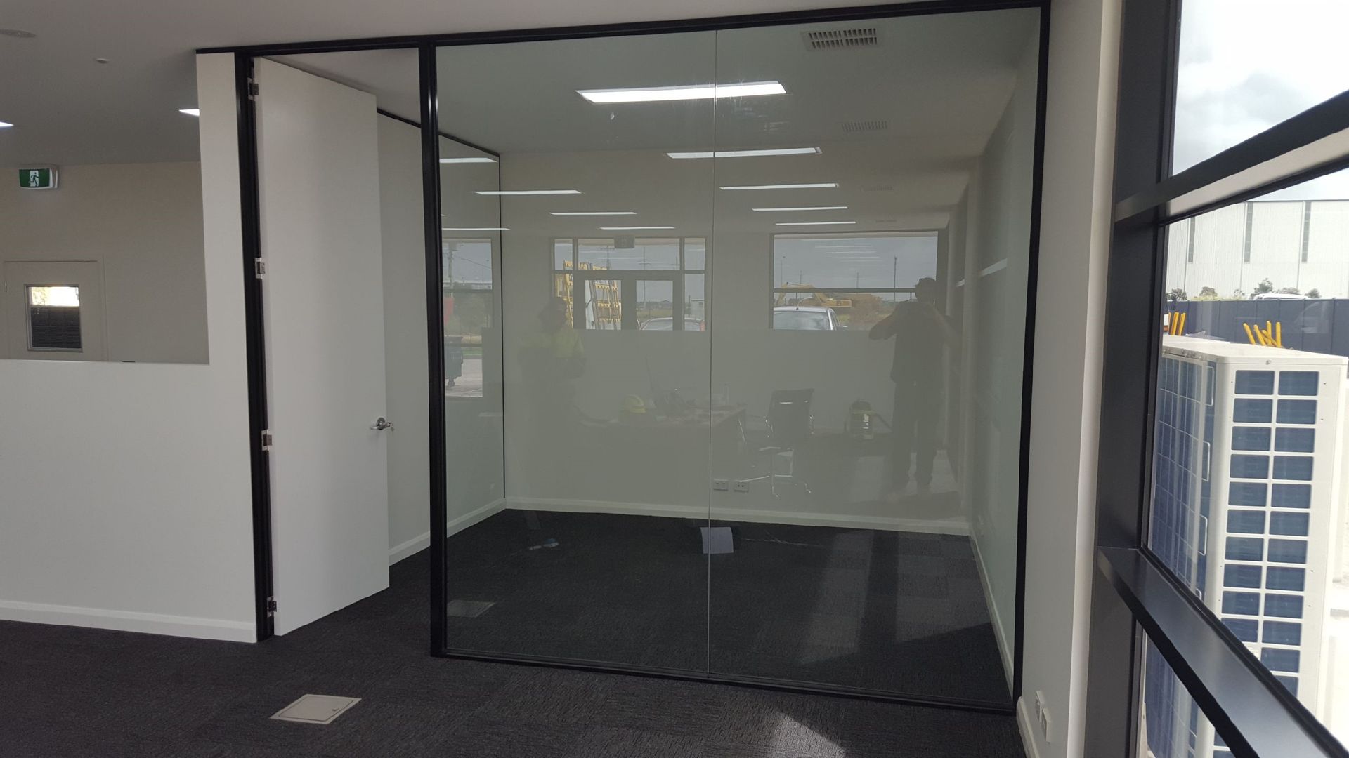 Glass-walled office space with black trim and dark floor, next to a white wall and a window with a city view.