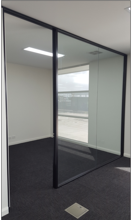 Glass-walled office interior with black trim, white walls, and dark carpet. A window offers an exterior view.