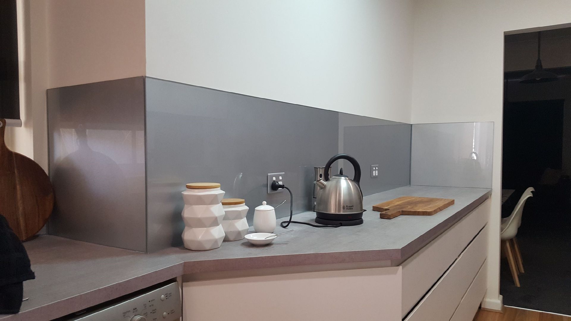 Kitchen with gray countertop, backsplash, white cabinets, kettle, jars, and wooden cutting board.