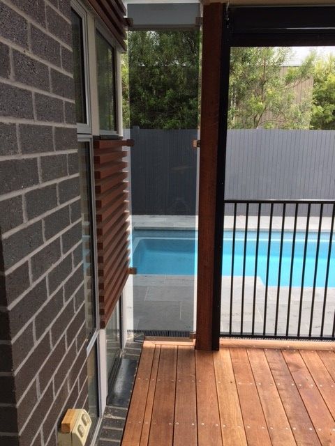 Wooden deck overlooking a pool and black metal fence, view from a brick building with wooden slats.