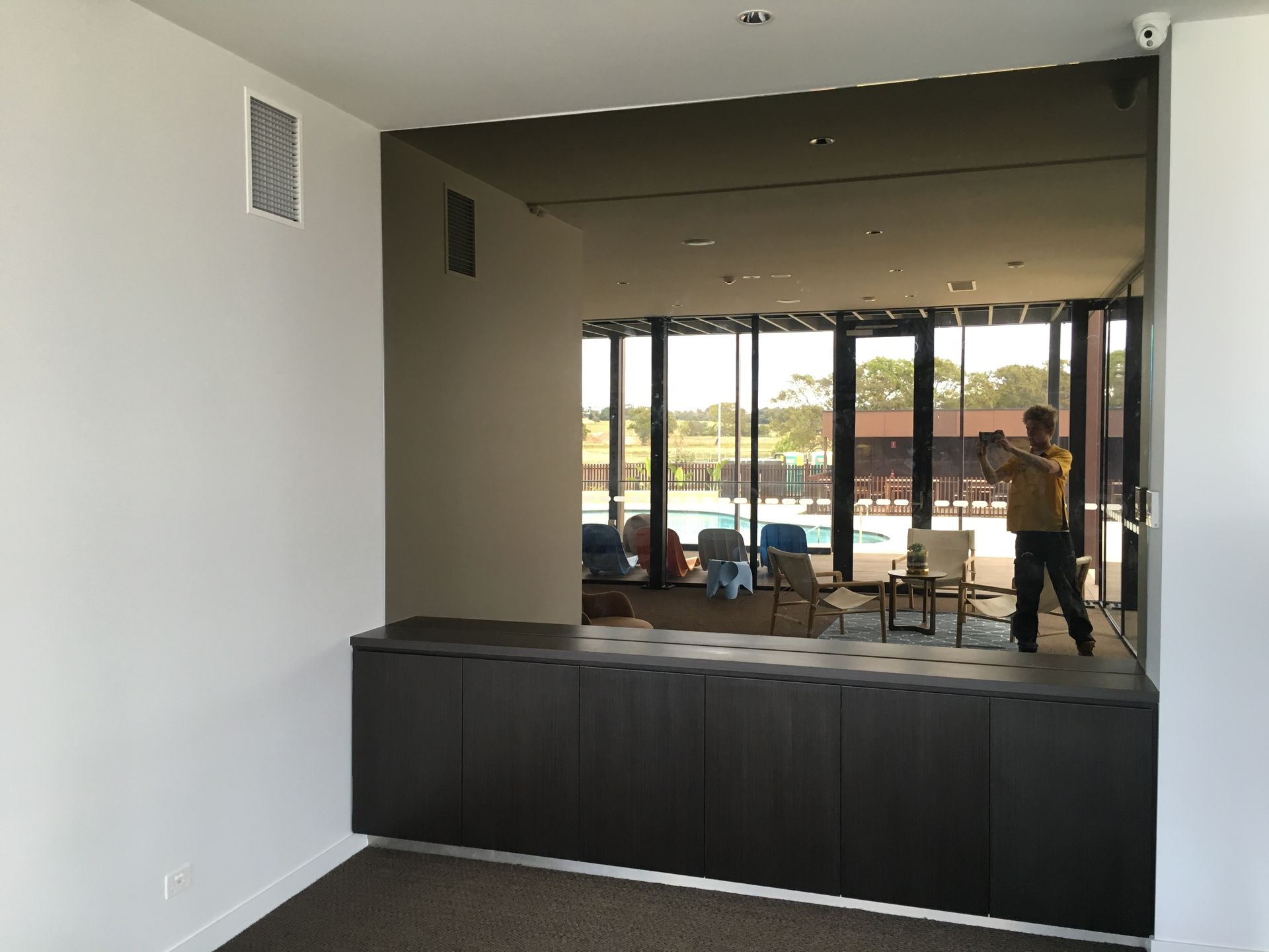 Mirror reflects a room with pool, man taking photo. Dark wood cabinets below, white walls.