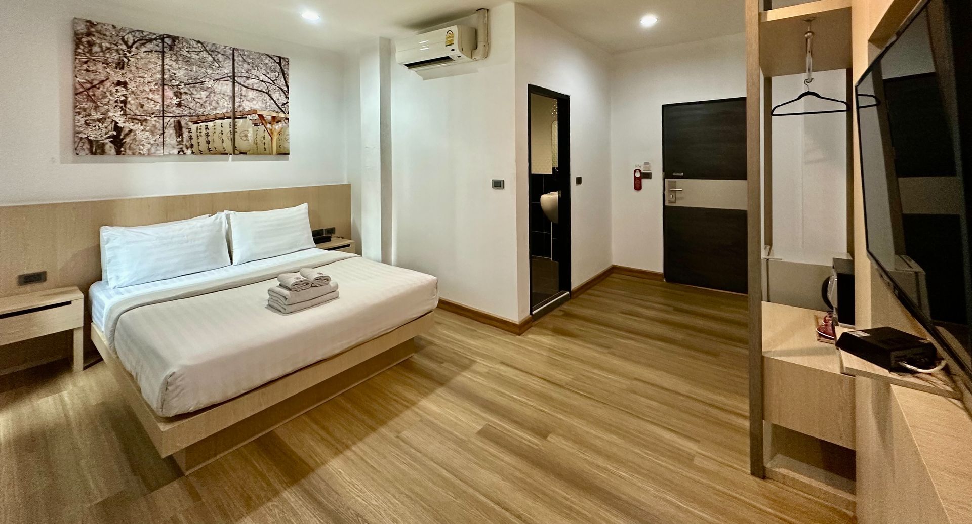 Hotel room with a king-sized bed, art, TV, closet, and a doorway to a bathroom. Wooden floors and neutral-colored walls.