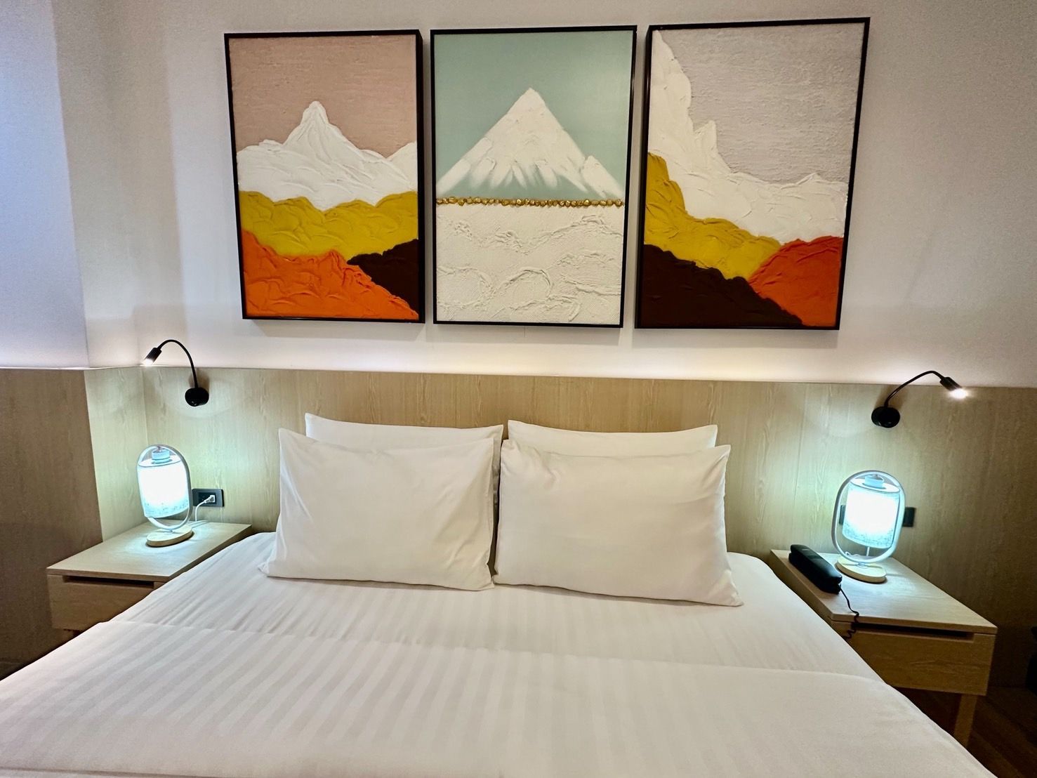 Bedroom with a made bed, nightstands, lamps, and three mountain paintings above the headboard.